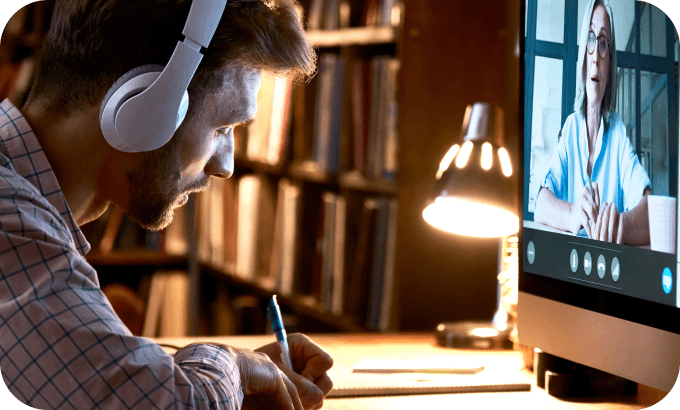 Man sitting at a desk at home, writing notes while in an online tutoring session on his laptop